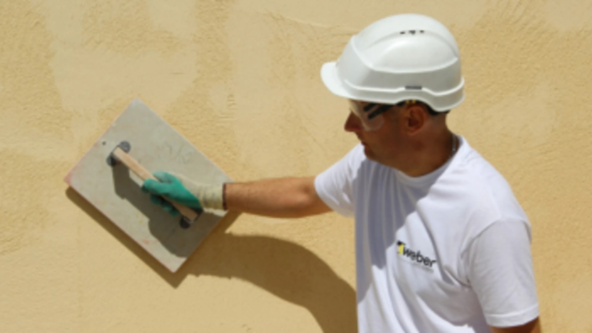 Muž v prilbe a ochranných okuliaroch aplikujúci žltú fasádnu omietku. Muž v prilbe a ochranných okuliaroch aplikujúci žltú fasádnu omietku.